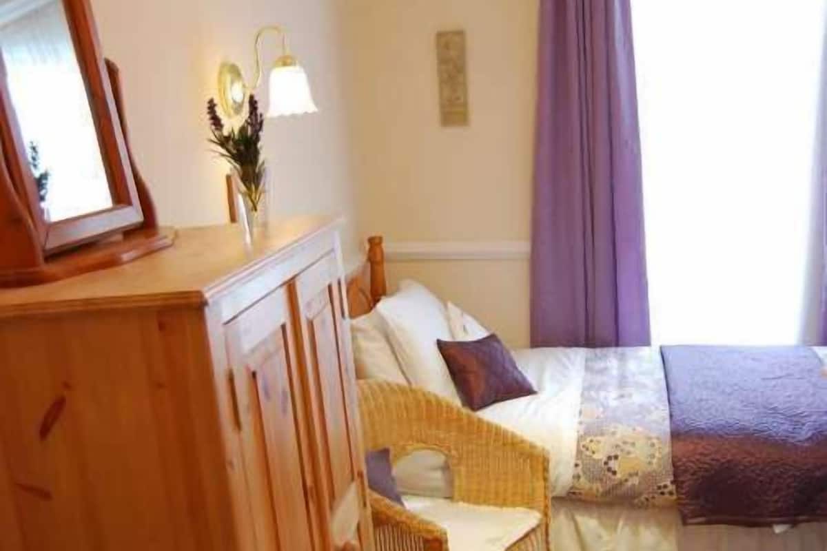 Ein Schlafzimmer mit einem Holfischschrank, einem Korbstuhl, einem Bett mit lila Decke und einem Fenster mit violetten Vorhängen.