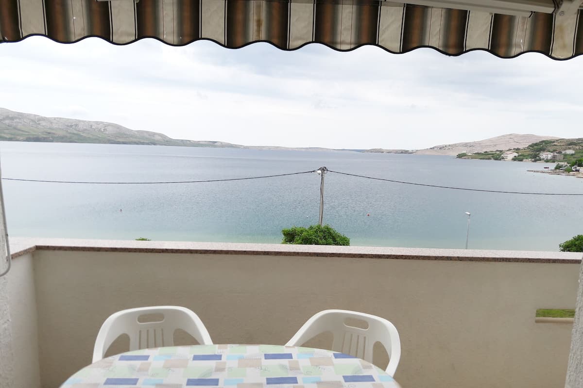 A balcony with a table and chairs, overlooking a calm body of water and distant hills.
