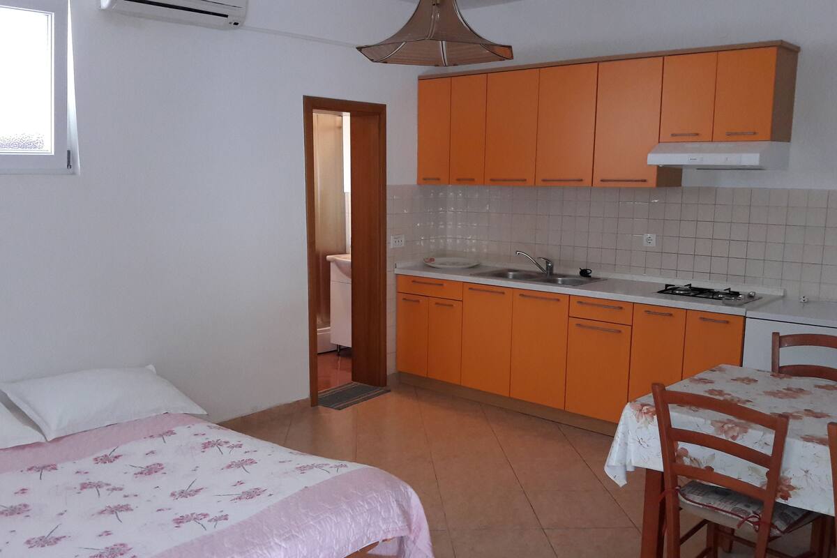 A compact kitchen with orange cabinets, a white countertop, and a sink. There is a dining table with a floral tablecloth and wooden chairs.