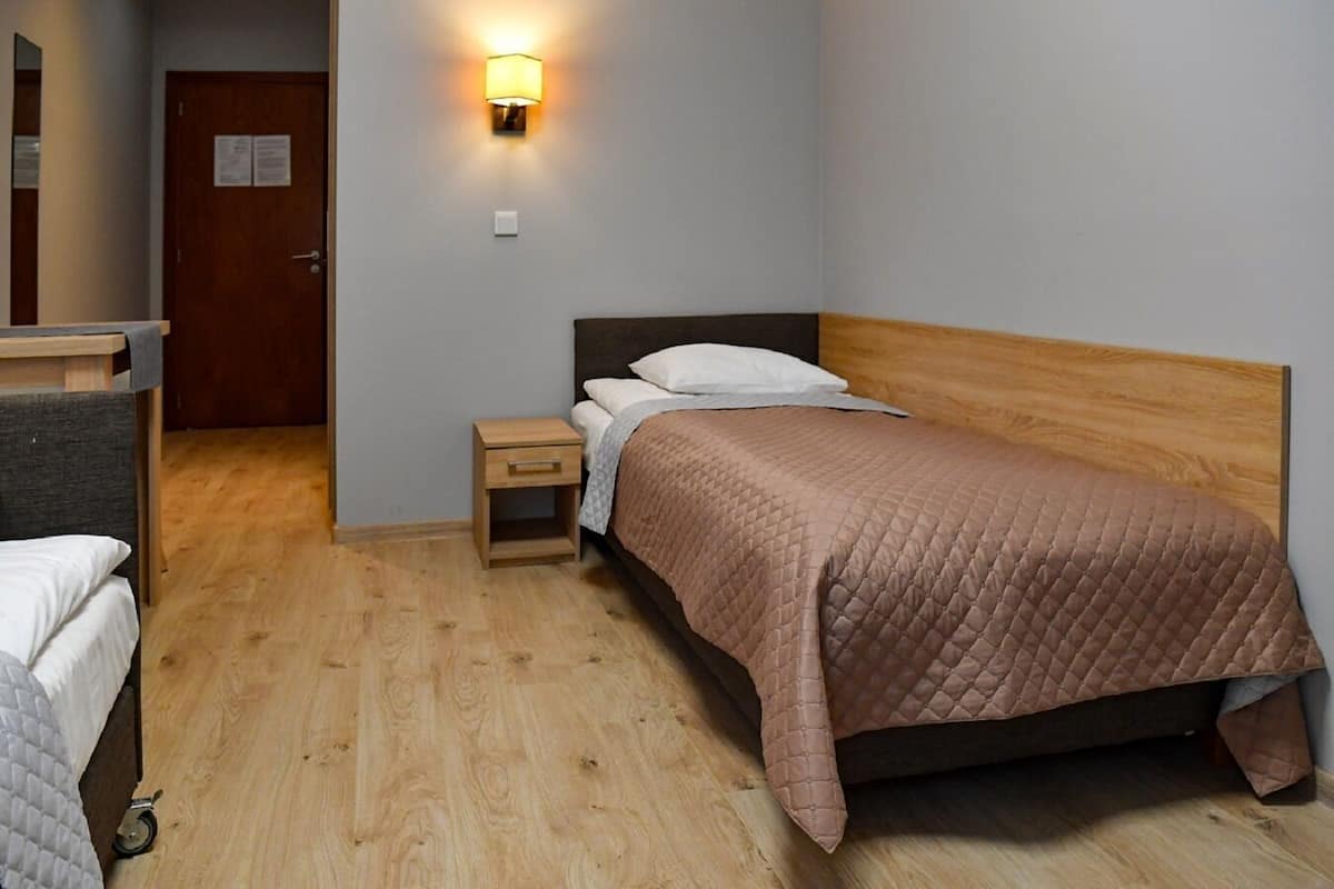 A single bed with a wooden headboard, a bedside table with a drawer, a wall-mounted light, and a door in the background.