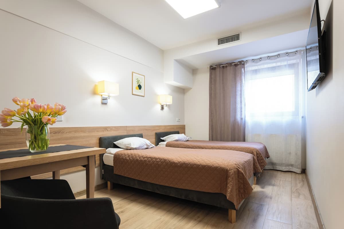 A hotel room with two beds, a desk, a chair, a vase with flowers, wall-mounted lamps, a flat-screen TV, and a window with curtains.