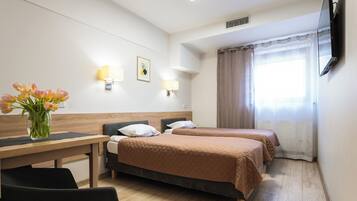 A hotel room with two beds, a desk, a chair, a vase with flowers, wall-mounted lamps, a flat-screen TV, and a window with curtains.