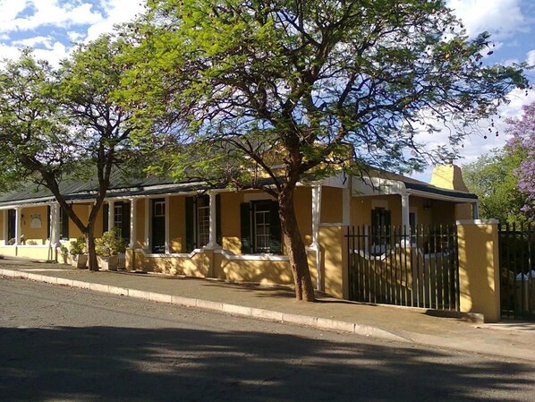 A yellow building with a gated entrance, a porch, and a tree in front. - Trackers Inn (Graaff-Reinet)