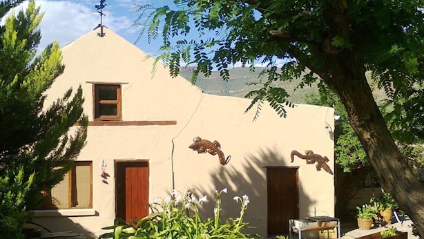 Il s’agit d’un bâtiment doté d’un toit en pente, de deux fenêtres et d’une porte. Des plantes et des arbres entourent le bâtiment.