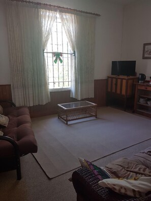 A living room with a sofa, a glass-top coffee table, a television on a wooden cabinet, and a window with curtains. - Trackers Inn (Graaff-Reinet)