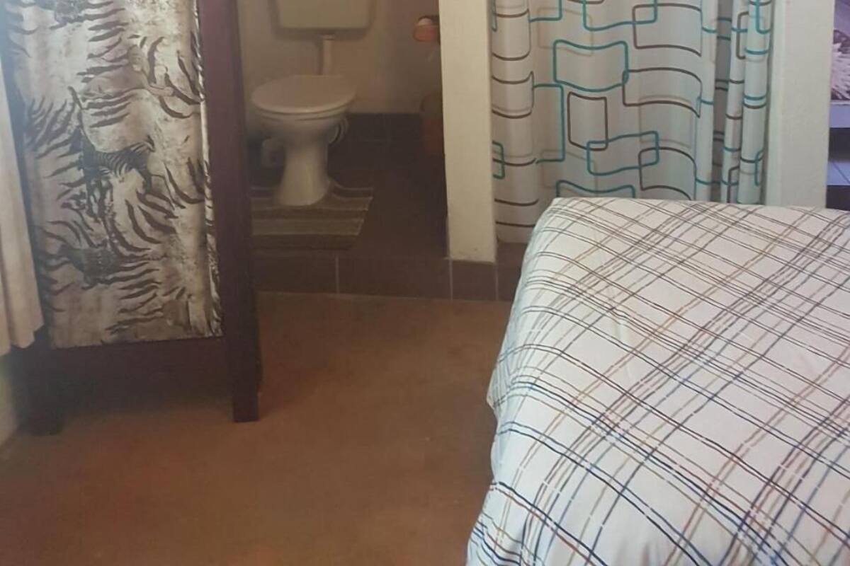 A small, dimly lit room with a wooden ceiling, a bed with a plaid bedspread, a patterned curtain, and a toilet visible through a doorway.