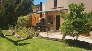 Un jardin avec un espace barbecue en pierre, une terrasse en bois et un petit arbre.