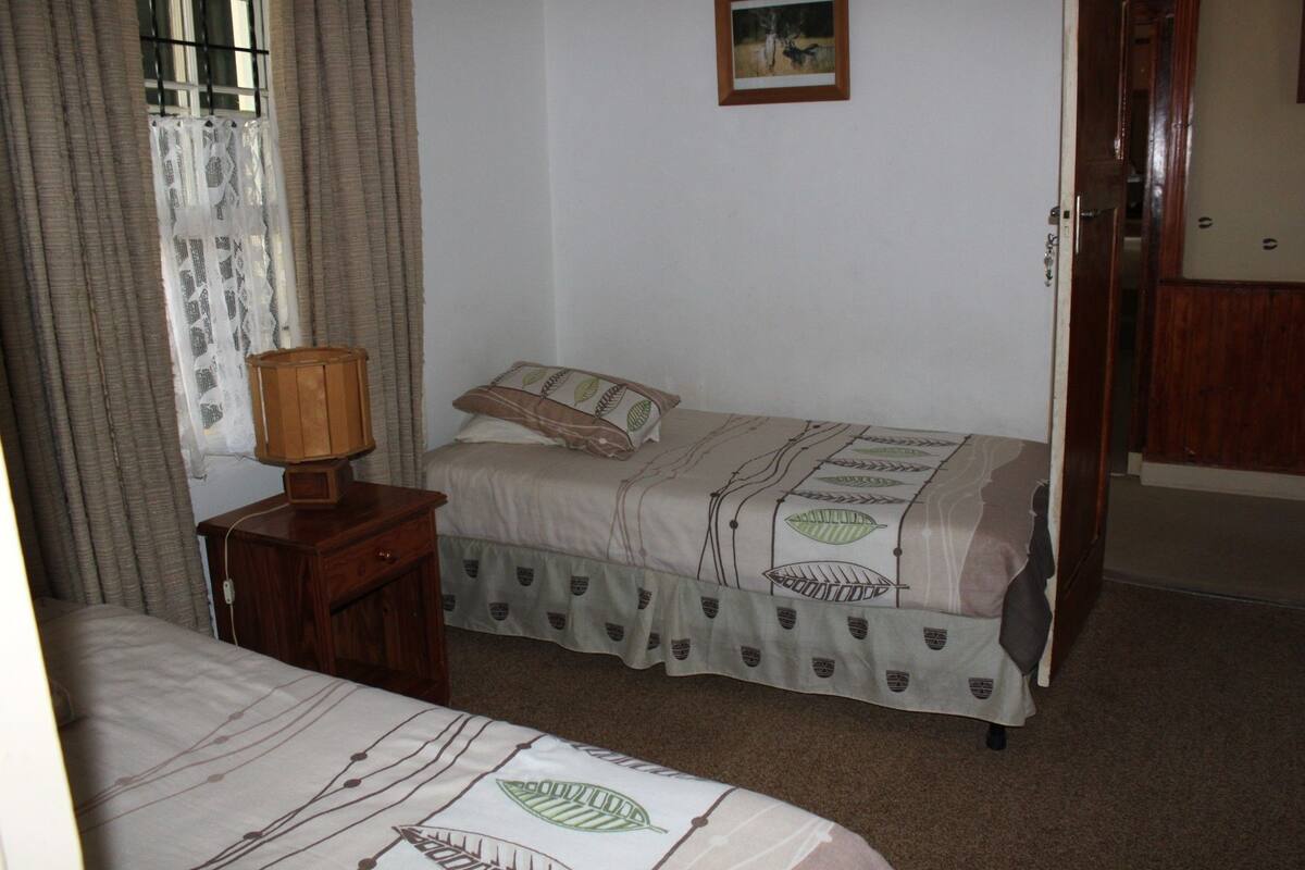 A bedroom with two single beds, a wooden nightstand with a lamp, a window with curtains, and a door.