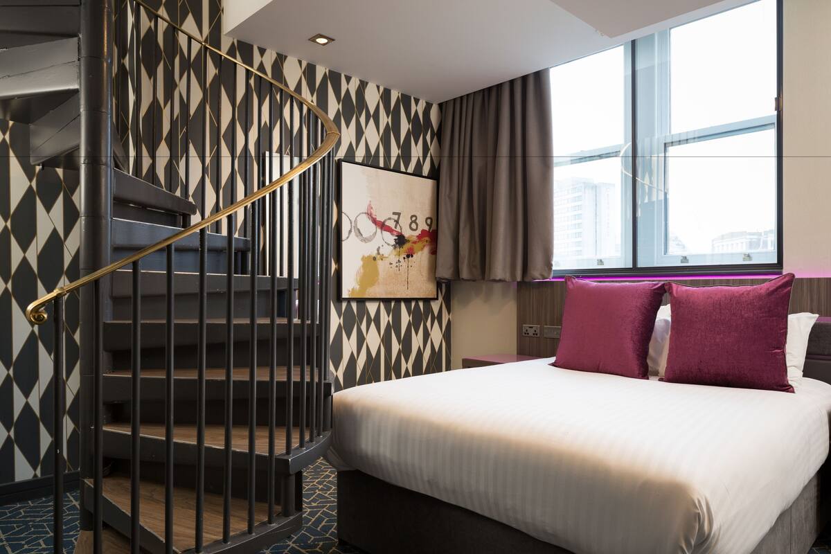A hotel room with a bed, a painting, a window, and a staircase.
