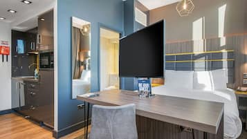 A modern hotel room with a bed, a desk, a chair, a flat-screen TV, and a 'Be Our Guest' sign.