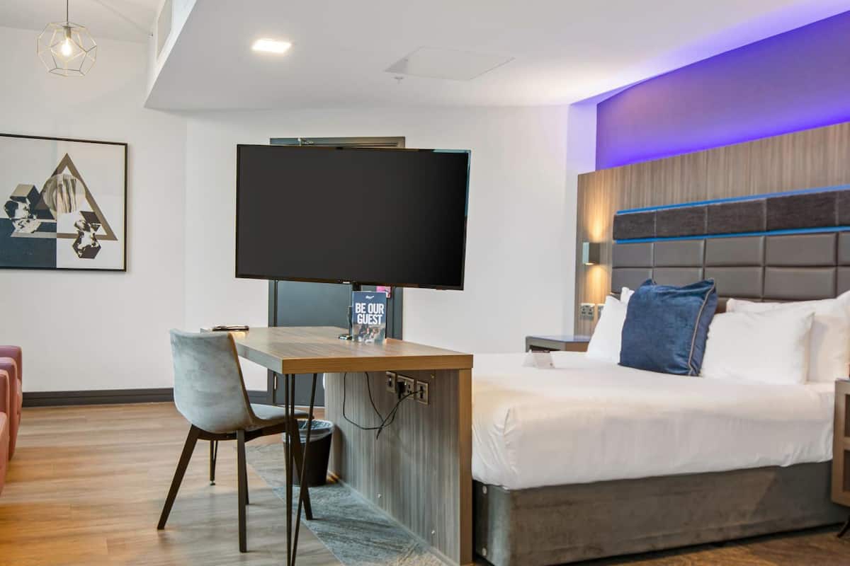 A modern hotel room with a bed, a desk, a chair, a flat-screen TV, and a 'BE OUR GUEST' sign.