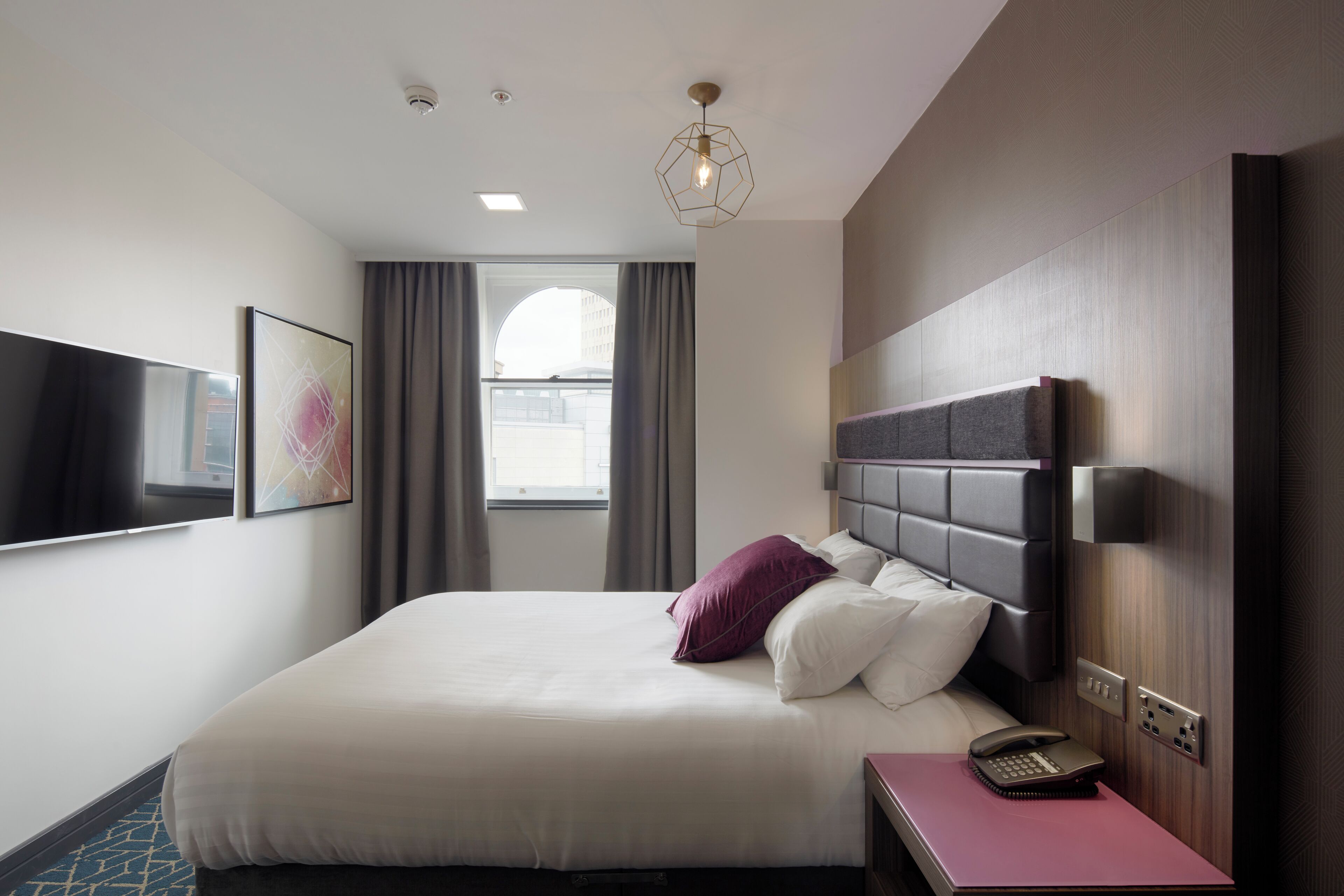Et moderne hotelværelse med en stor seng, et fladskærms-tv, et skrivebord med telefon og et vindue med gardiner.