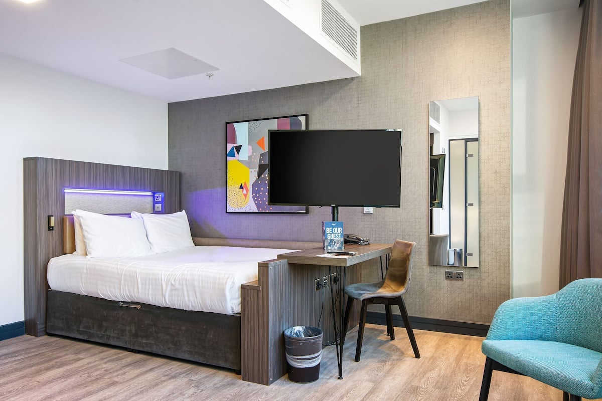 A hotel room with a bed, desk, chair, TV, and a trash bin.
