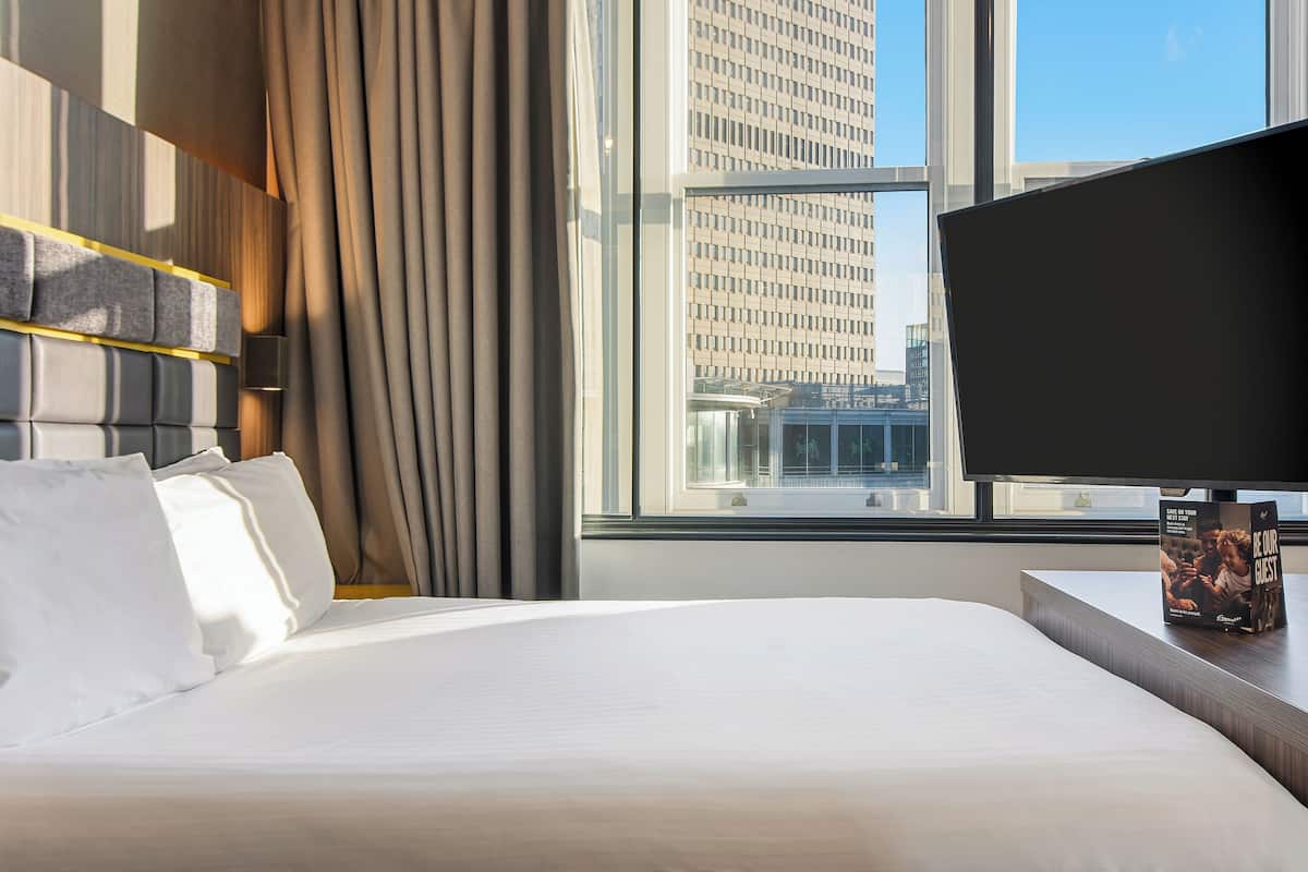 A hotel room with a large bed, a flat-screen TV, a window with a city view, and a bedside table with a magazine.