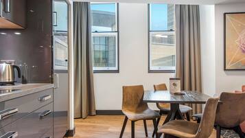 Ein modernes Hotelzimmer mit Küchenzeile, einem Esstisch mit Stühlen und großen Fenstern mit Blick auf die Stadt.