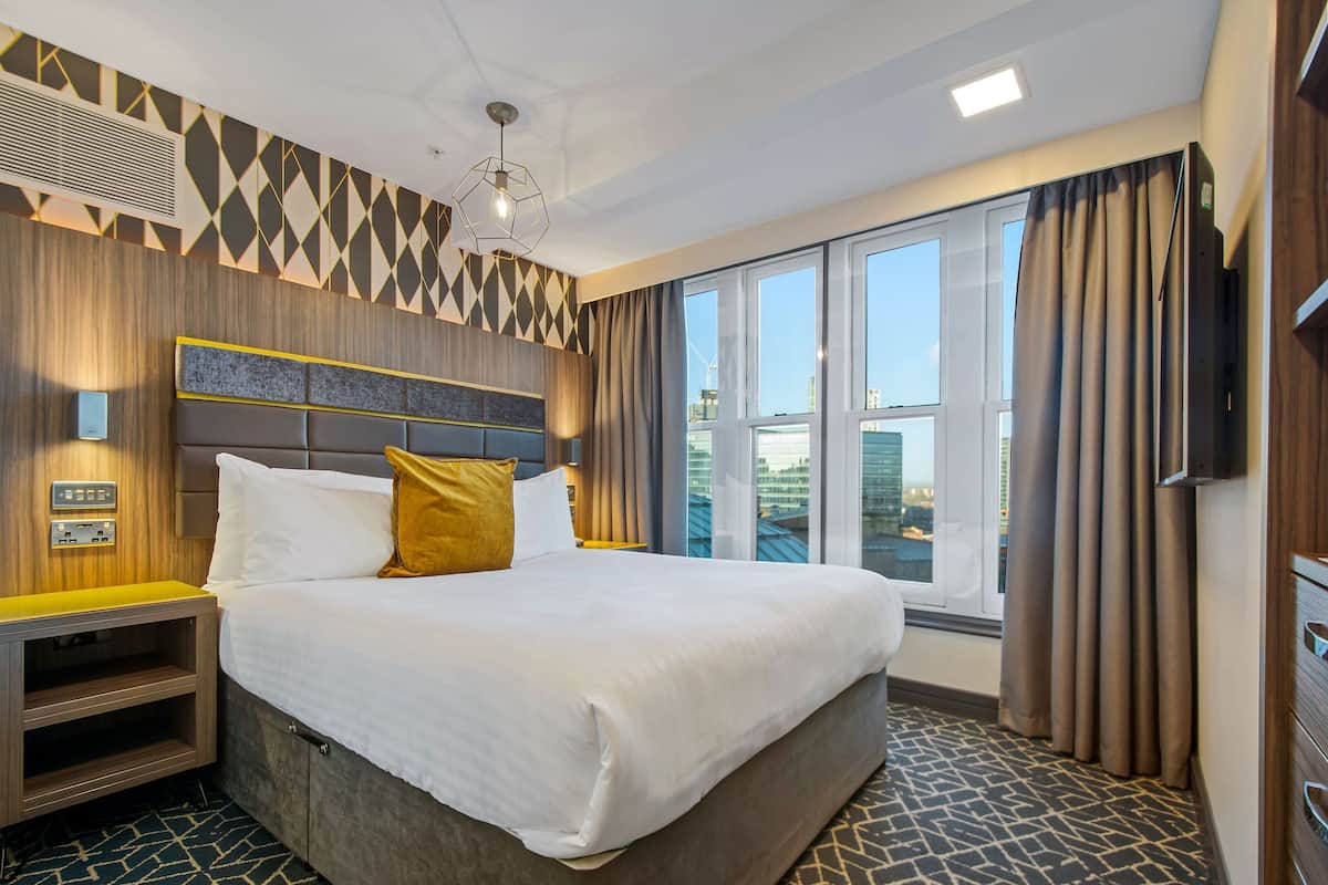 A modern hotel room with a large bed, a nightstand, a TV, and a window with curtains.