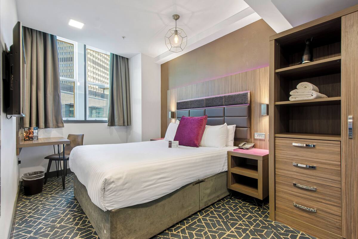 A hotel room with a bed, desk, chair, wardrobe, and a view of buildings through the window.