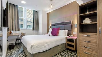 A hotel room with a bed, desk, chair, wardrobe, and a view of buildings through the window.