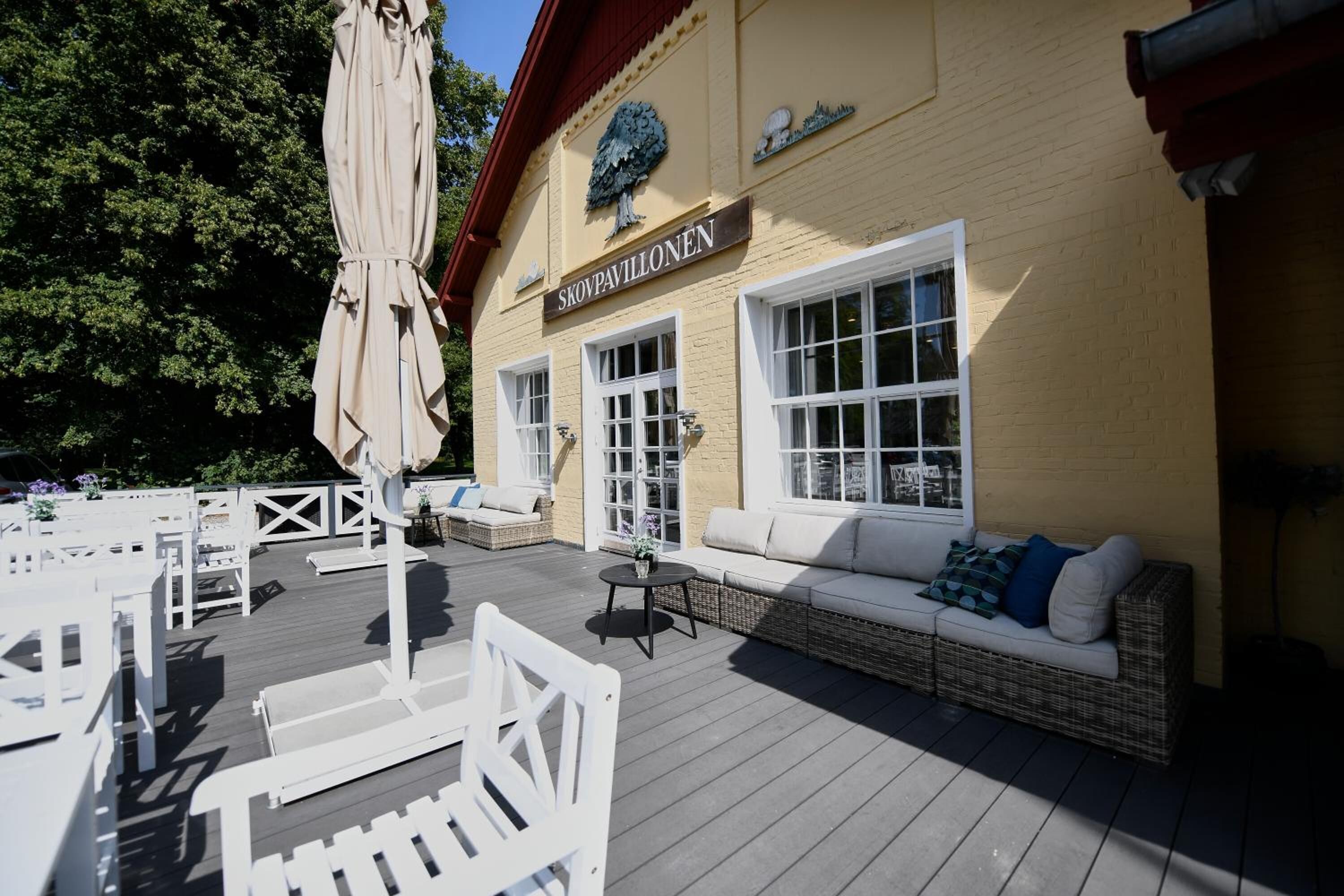 Una terraza de madera con mobiliario exterior, incluyendo sillas blancas y un sofá, y un edificio con un letrero que dice "SKOVPAVILLONEN".