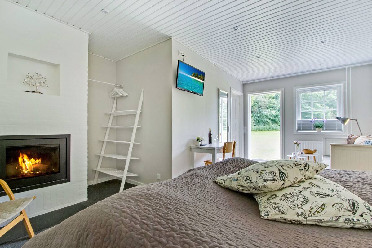 Une chambre avec une cheminée, un lit agrémenté d’oreillers à motifs, un téléviseur fixé au mur et une vue sur la verdure à l’extérieur.