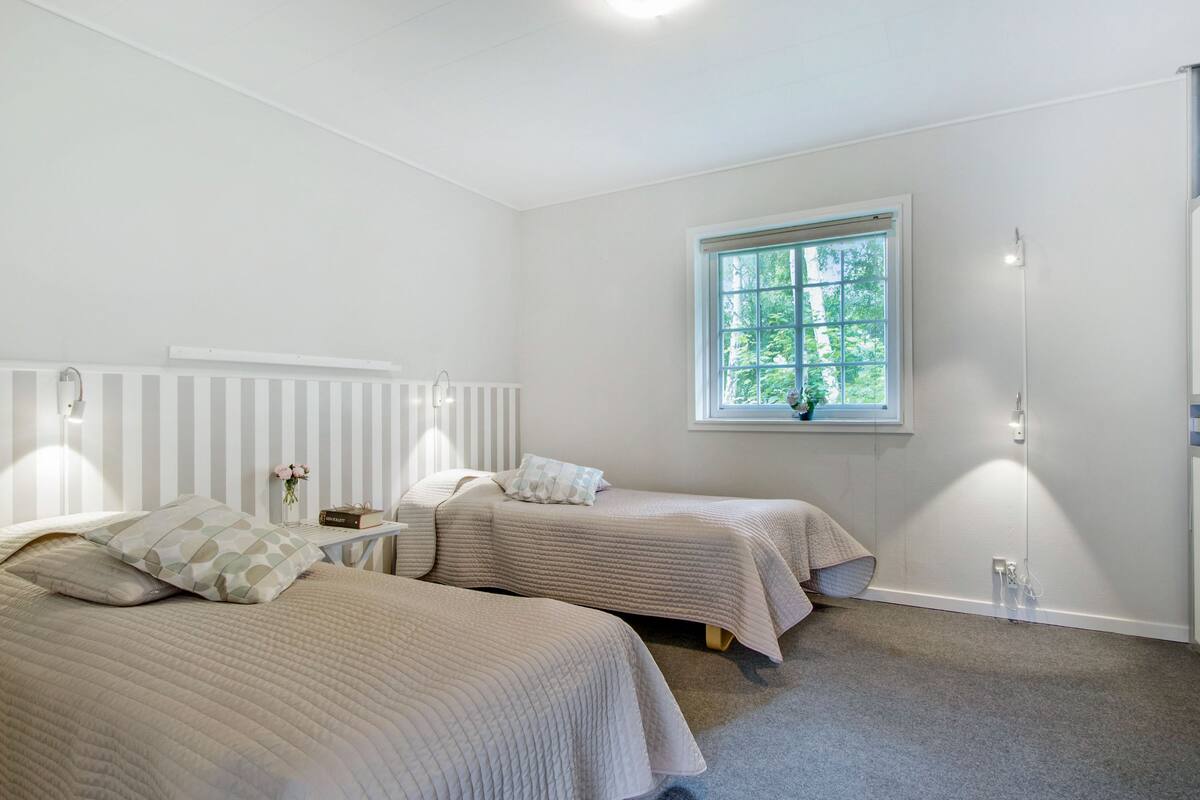 Une chambre avec deux lits, une fenêtre donnant sur un espace verdoyant, un radiateur et une table de chevet avec une lampe.