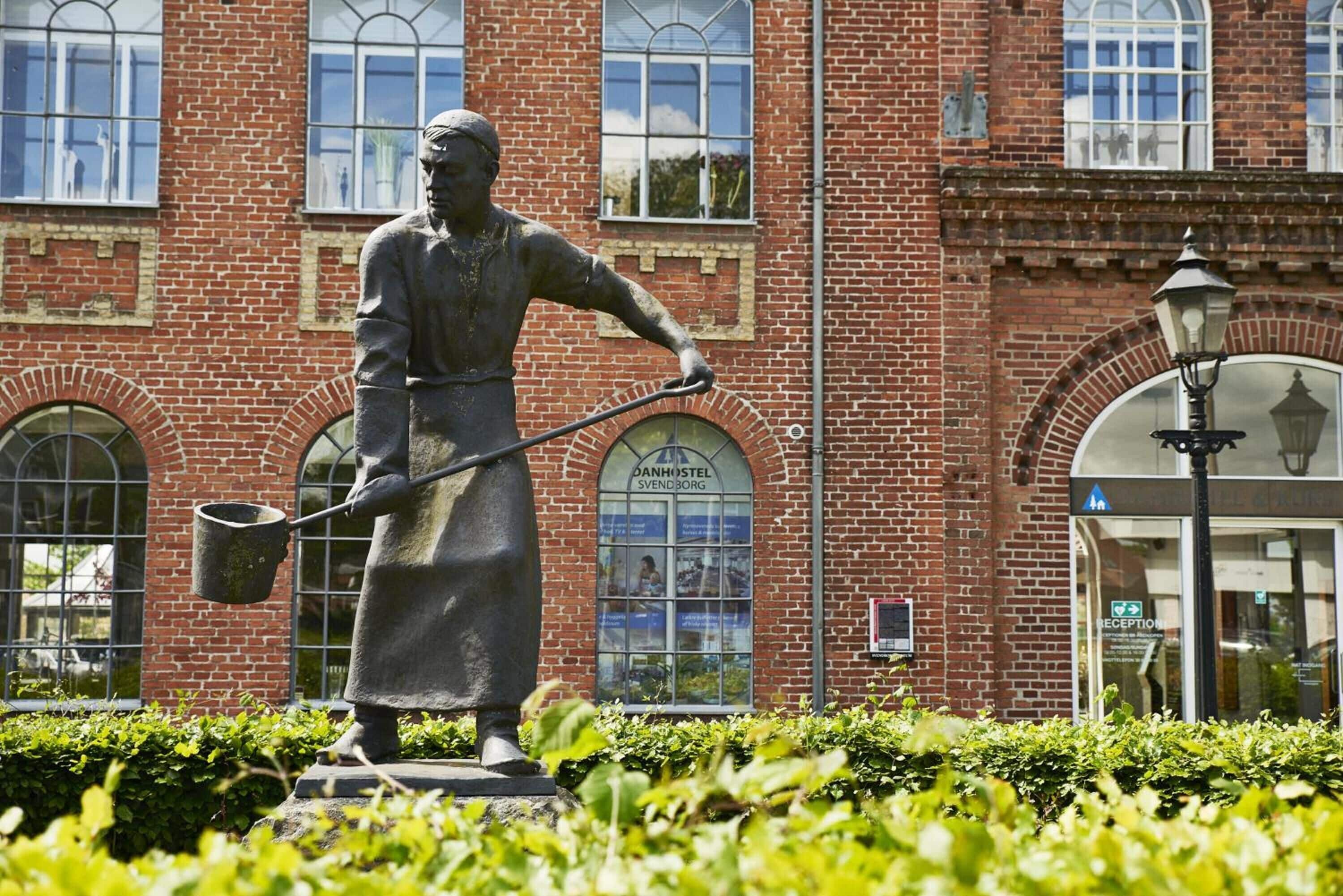 A statue of a man holding a shovel in front of a brick building with arched windows and a sign that reads "DANHOSTEL SVENDBORG".