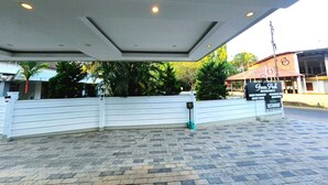 A gated entrance to a property with a paved walkway, a signboard advertising amenities, and a building with a red roof in the background. - Green Park residency (Kannur)