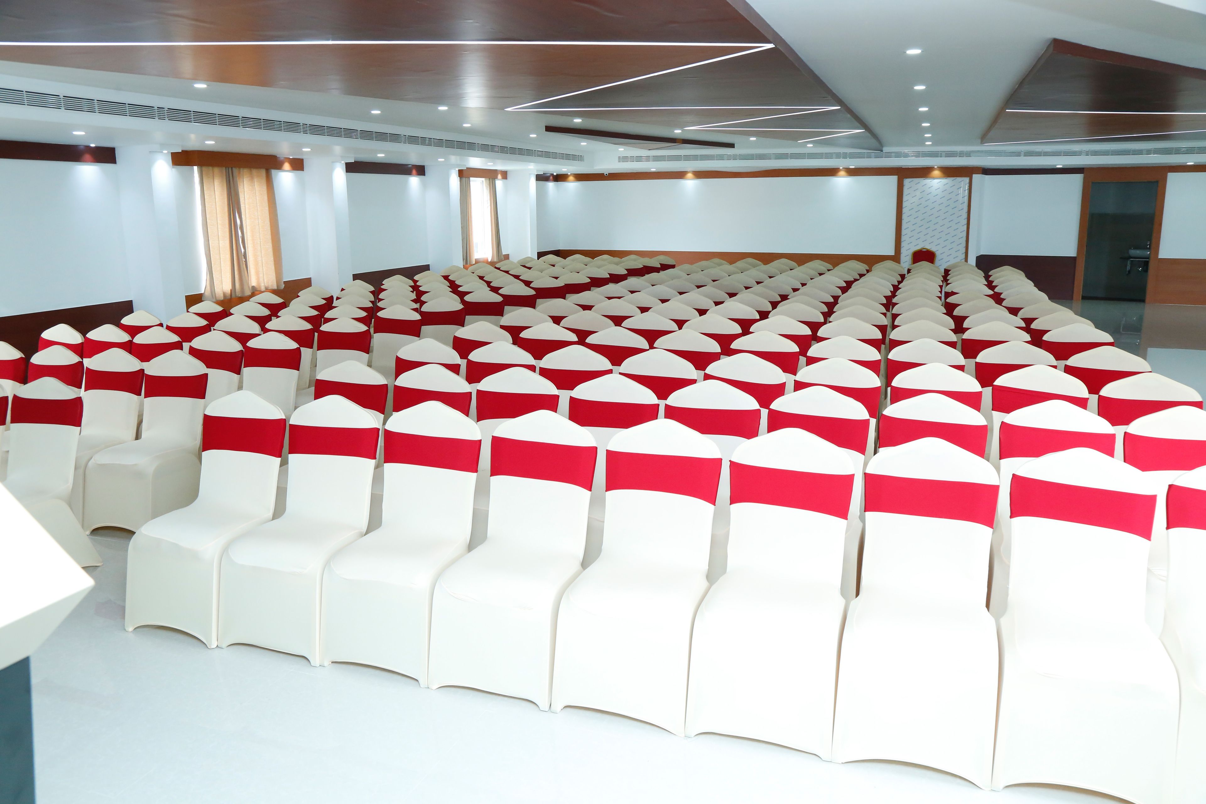 A large conference room with rows of white chairs with red sashes, a wooden ceiling, and a large window with curtains.