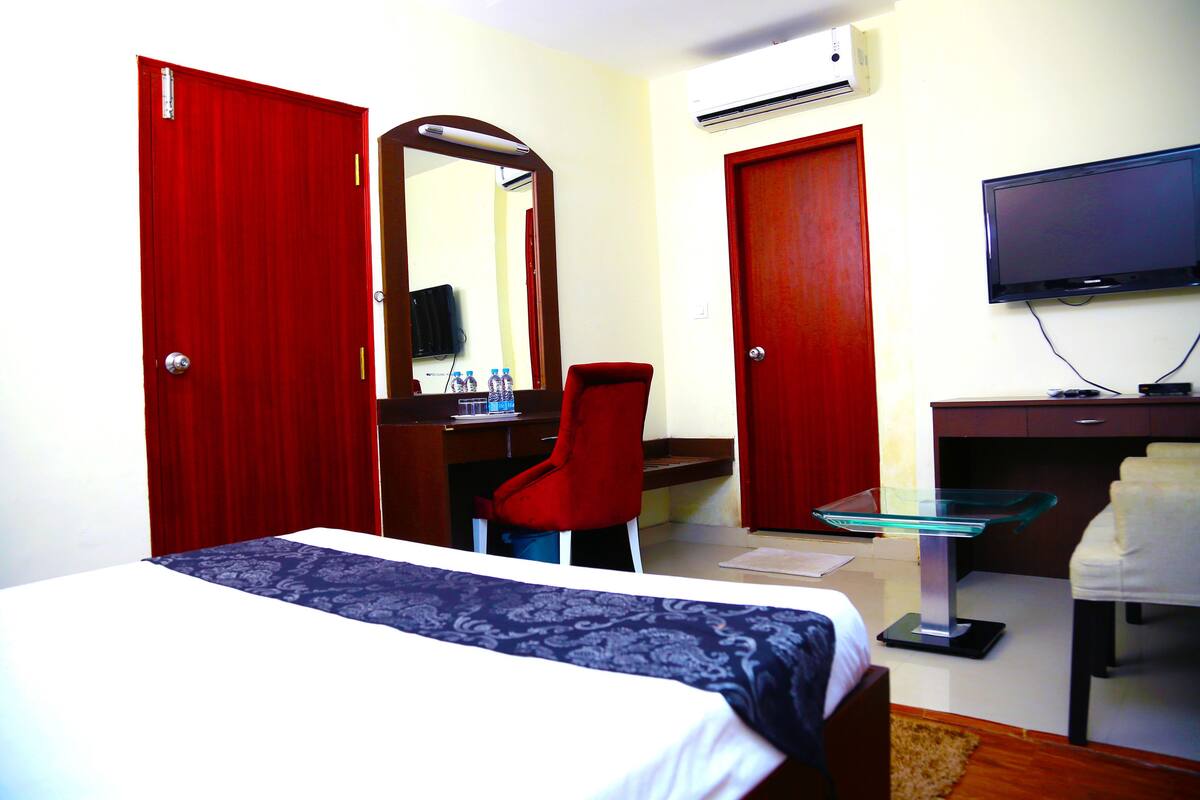 A hotel room with a bed, a desk with a chair, a TV, and two red doors.