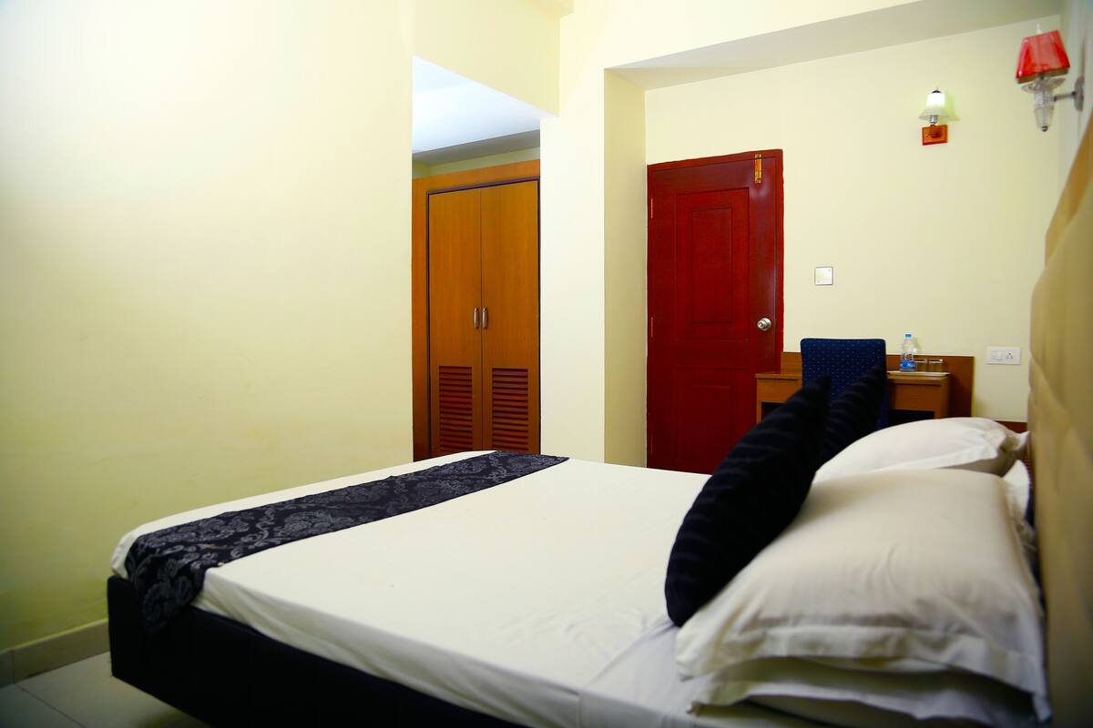 A hotel room with a bed, a nightstand, a red door, and a small table with a lamp.