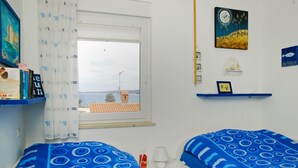 A room with two beds, a window with a view of a distant building, and a shelf with decorative items. - Segundo al Mar Apartments (Medulin)