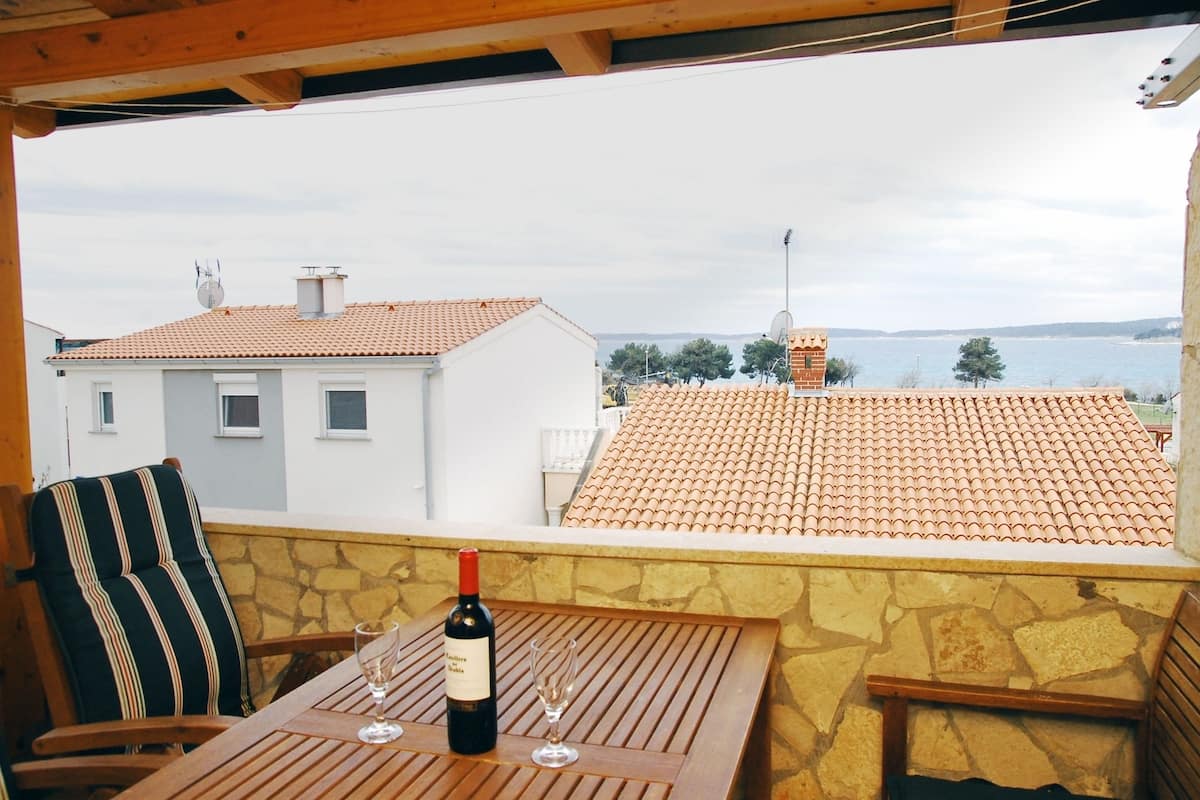 A terrace with a table, a bottle of wine, and a glass. The terrace overlooks a body of water and houses with red-tiled roofs.