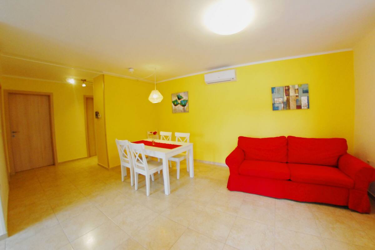 Une pièce aux murs jaunes, avec un canapé rouge, une table à manger blanche et des chaises, ainsi qu’un luminaire suspendu.