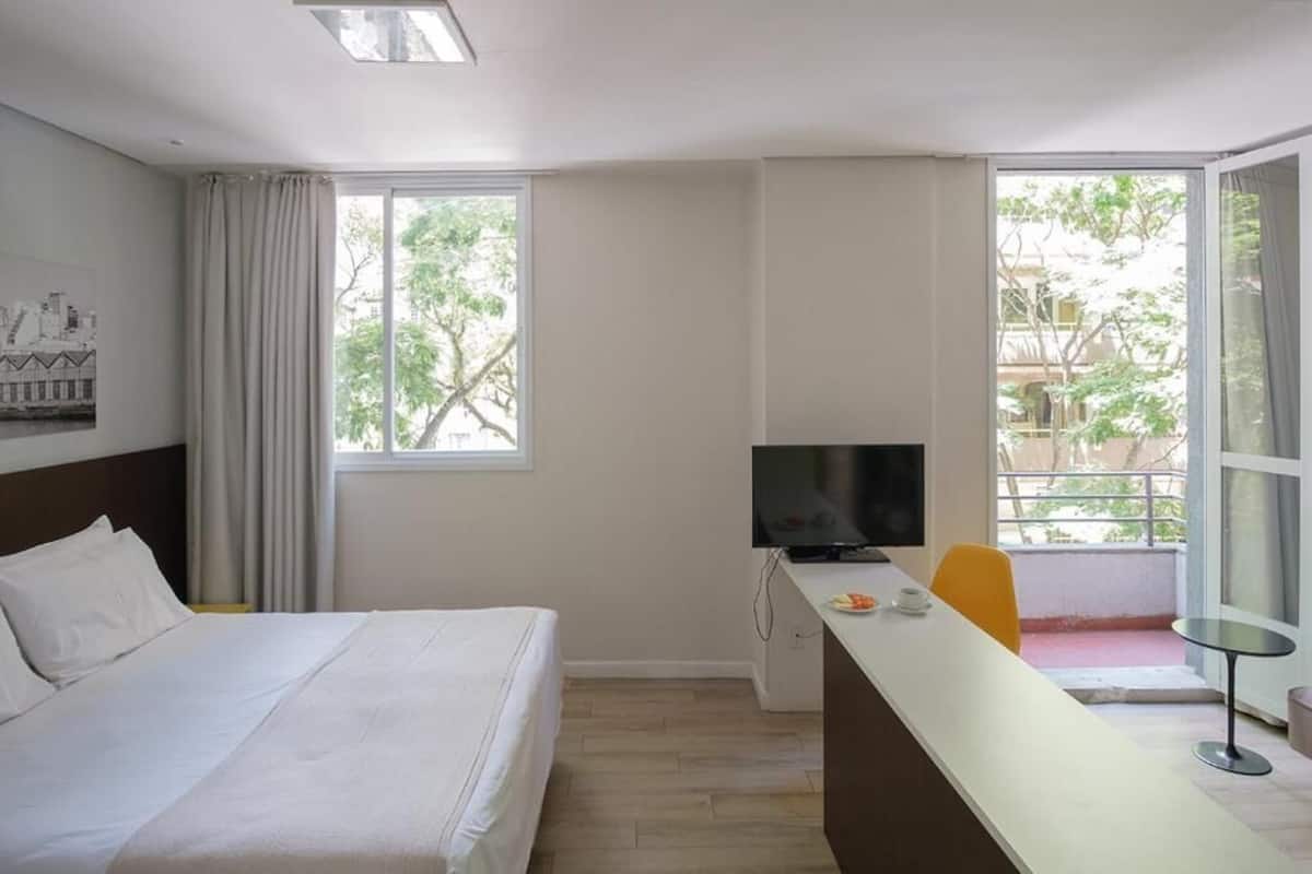 Quarto de hotel com cama, escrivaninha, televisão e varanda com vista para árvores.