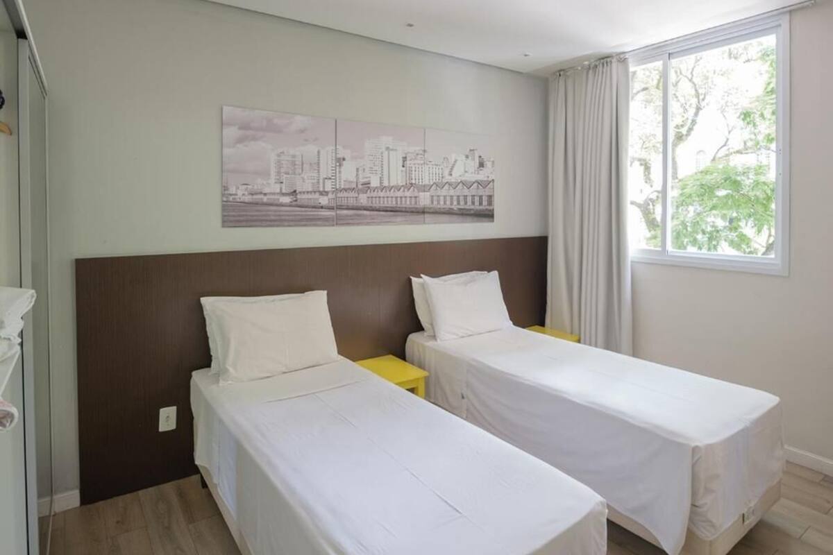 Quarto de hotel com duas camas de solteiro, uma pintura com vista urbana na parede e uma janela com cortinas.