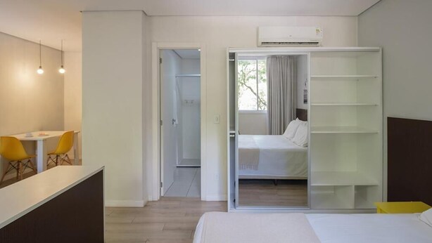 A modern hotel room with a bed, a desk with chairs, a wardrobe, and an air conditioning unit. - Açores Flat Design (Porto Alegre)