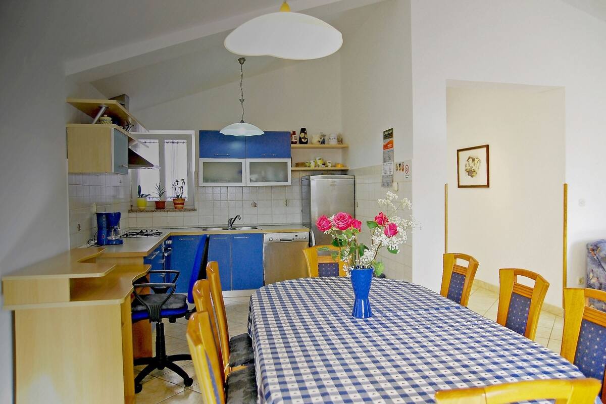 A compact kitchen with blue cabinets, a dining table with a checkered tablecloth, and a vase with flowers.