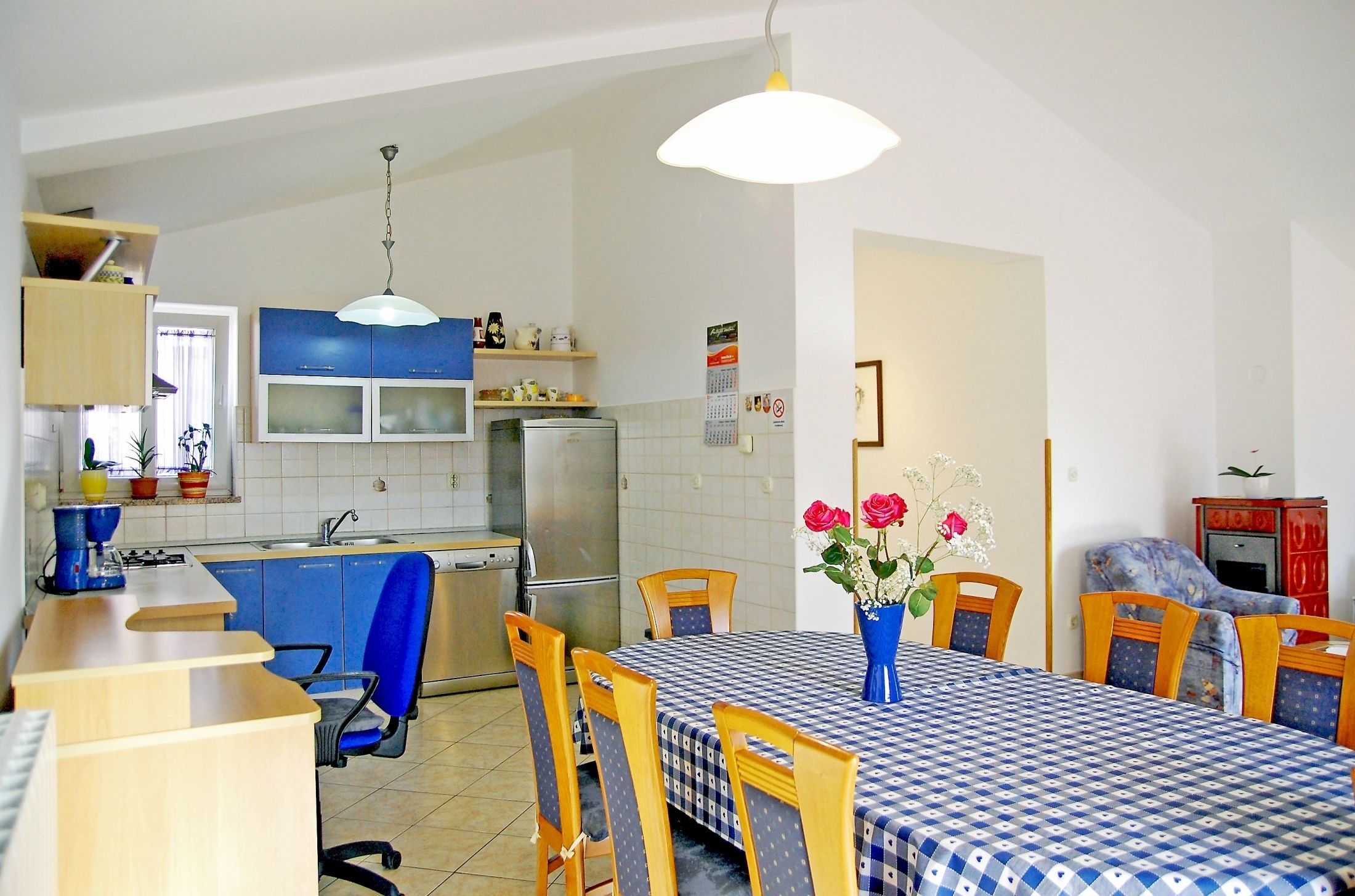 A compact kitchen with a dining area, featuring a blue and white checkered tablecloth, wooden chairs, and a vase with flowers.
