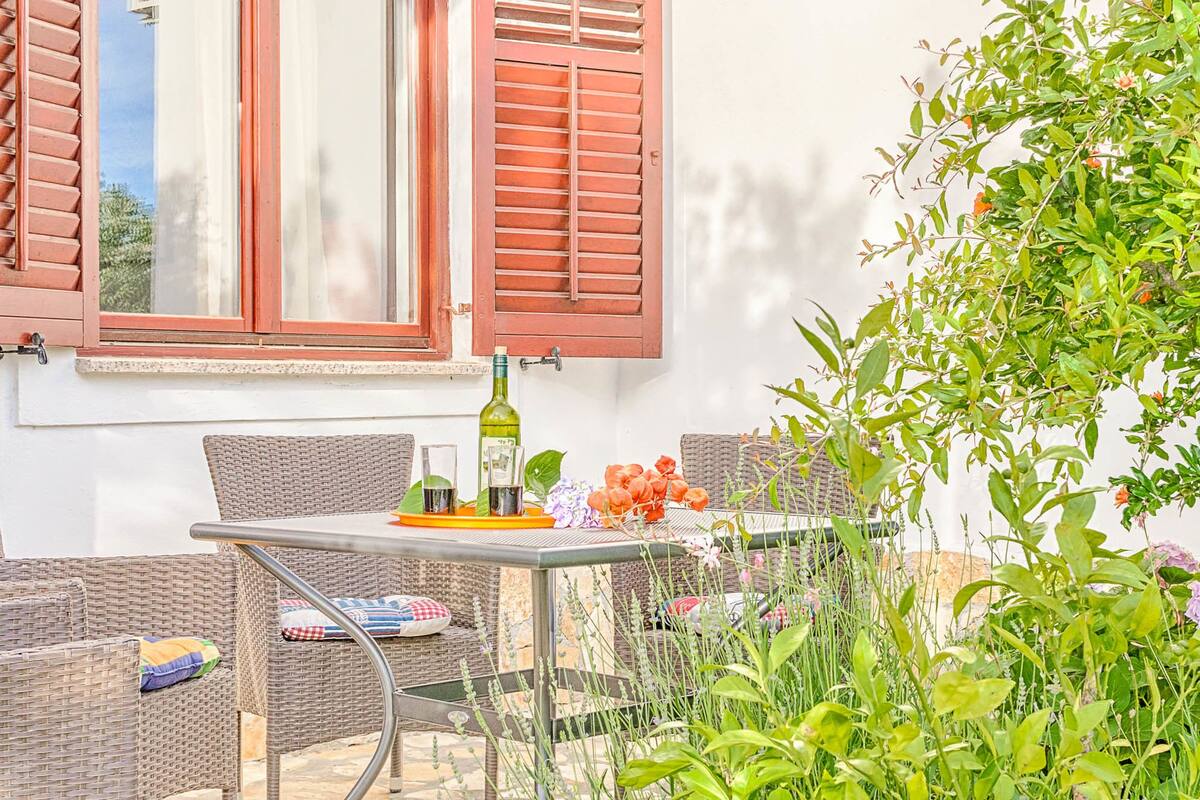 A patio with wicker furniture, a table with a bottle and glasses, and a view of a building with red shutters.