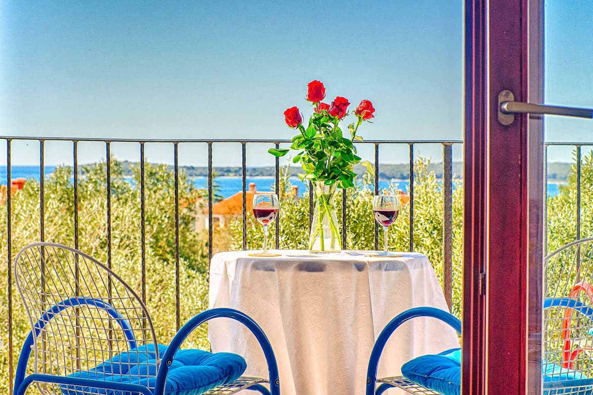 A balcony with a table set for two, featuring a vase with red flowers and two wine glasses.
