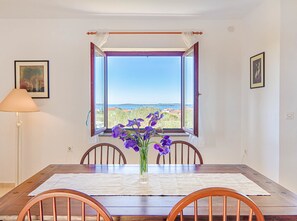 A dining table with chairs, a vase with purple flowers, and a view of the sea through an open window.