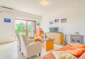 A spacious living area with a sofa, a coffee table, a TV, and a cabinet. There is a window with a view of trees and a door leading outside.