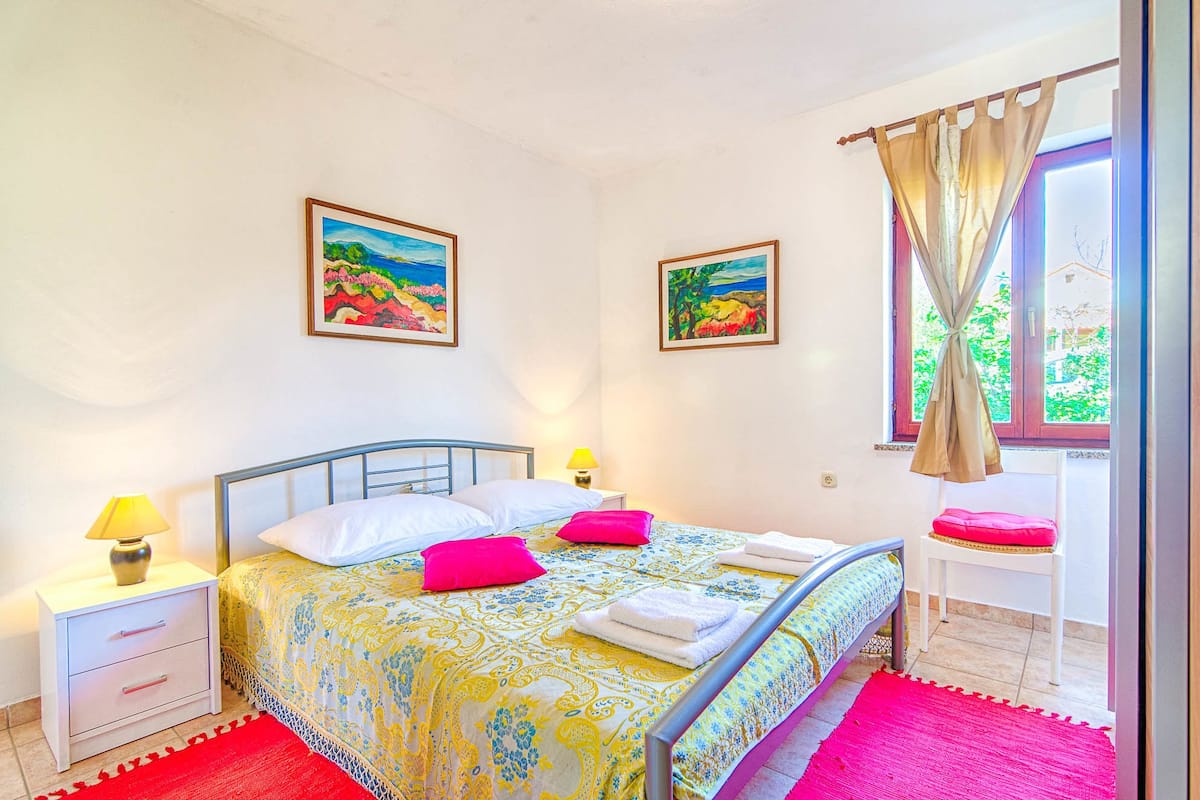 A bedroom with a bed, two nightstands, a window with curtains, and two paintings on the wall.