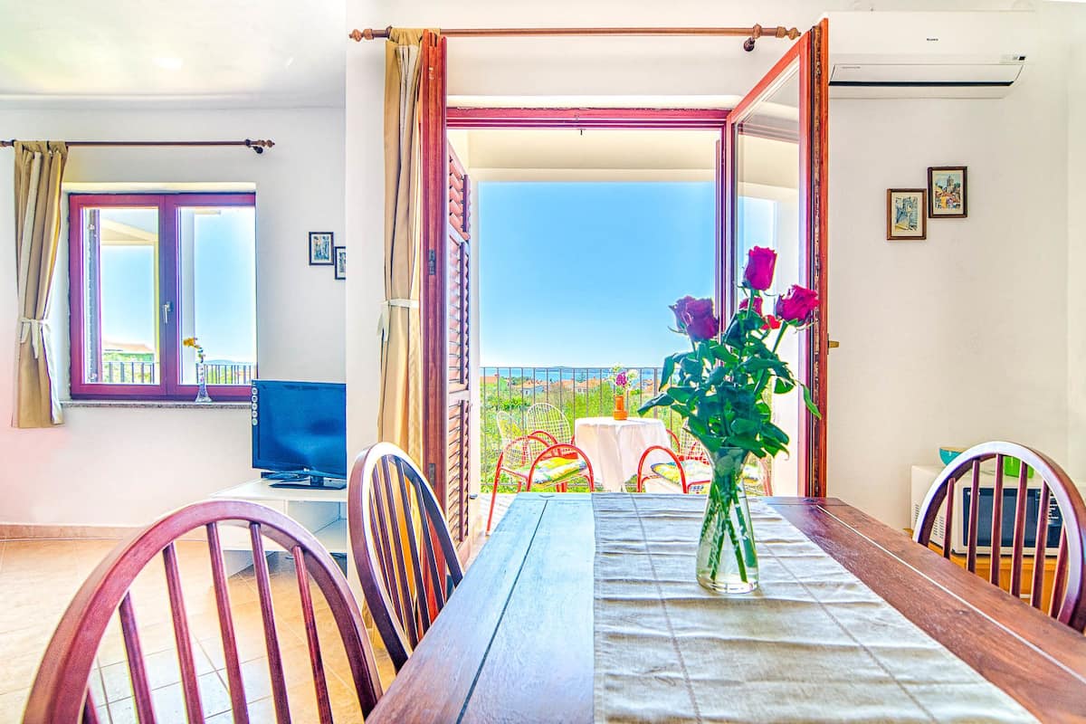 A dining area with a table, chairs, a vase with flowers, and a view of the outdoors.