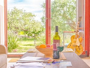 A room with a large window, a guitar, a bottle of wine, a bowl of fruit, and some magazines on a table.