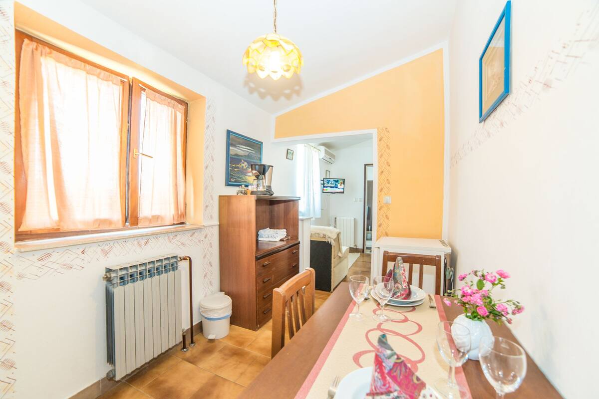 Un coin repas avec une table en bois dressée pour deux personnes, un radiateur, une fenêtre avec des rideaux et un téléviseur en arrière-plan.