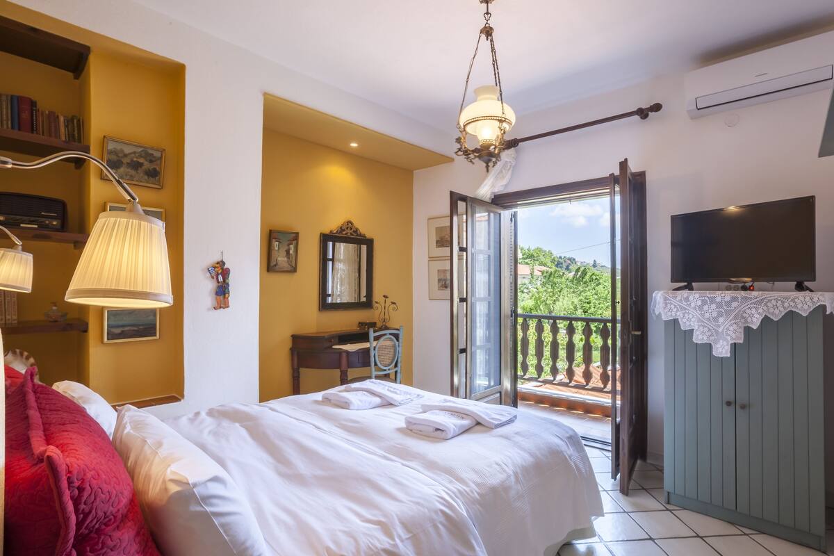 A hotel room with a bed, a desk, a chair, a television, and a balcony with a view of greenery.