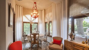 A room with a stone floor, a dining table with chairs, a sofa with a red cushion, a wooden cabinet with decorative items, and large windows with curtains. - Alterra Vita (Sithonia)