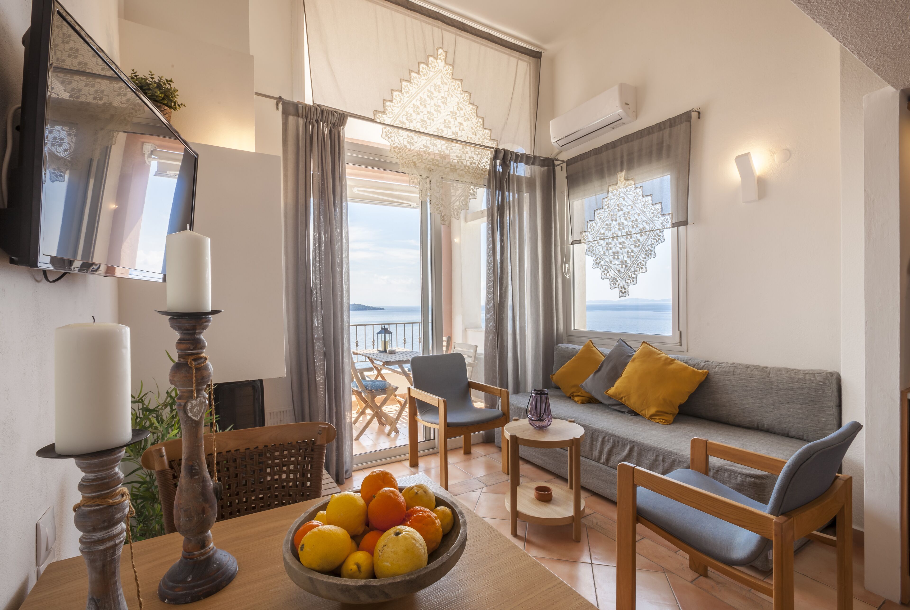 Ein gemütliches Wohnzimmer mit einer Couch, Sesseln, einem Tisch mit Früchten, einer Kerze und Blick auf das Meer.