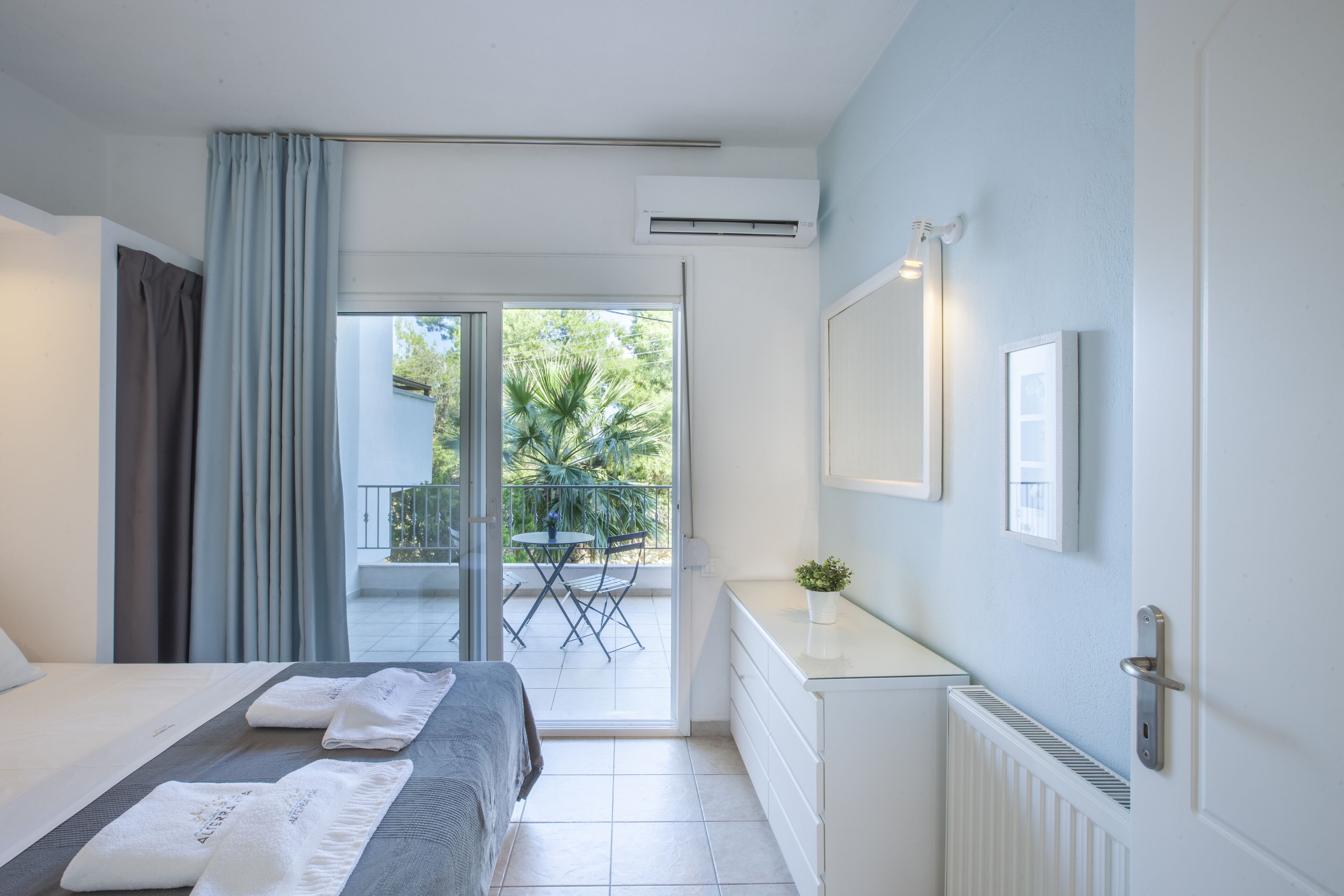 Quarto de hotel com cama, uma pequena escrivaninha, uma planta em vaso e vista para uma varanda com mesa e cadeiras.